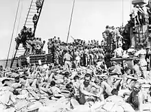 Black and white photo of a large number of men crowded together on the deck of a ship. Those in the foreground are sunbaking with their shirts off. Those in the background are standing with their backs to the camera. Some of the men in the background have climbed the ship's rigging or onto structures on the deck.